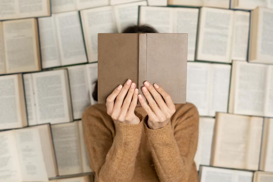 A girl in a sweater lying on a stack of open books, hiding her face with one book